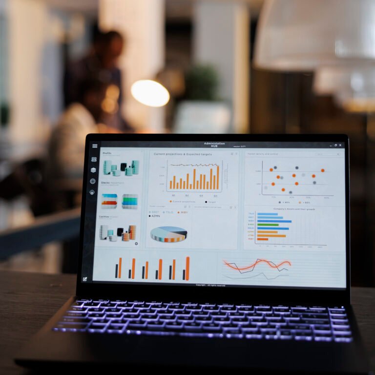 Laptop computer with marketing statistics graphs on screen, standing on table in startup office, coworkers discussing company strategy in background. Employees working overtime in startup office