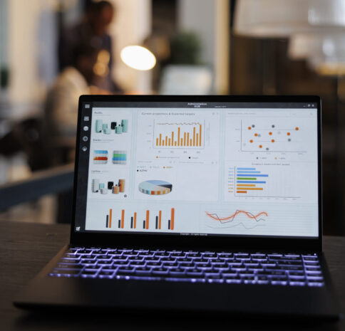 Laptop computer with marketing statistics graphs on screen, standing on table in startup office, coworkers discussing company strategy in background. Employees working overtime in startup office
